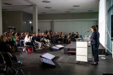 Blick in den Veranstaltungs-Saal: Rednerpult, dahinter die Bühne mit Podium und Projektionsfläche, davor Screens mit Zeitmanagement, Foliensätzen sowie ausgewählten Fragen aus dem Chat der hybriden Konferenz. (Bildnachweis: 18. Internationales Bodensee-Symposium, 2024 © ICOM Deutschland, Foto: Nicolas Bühringer)